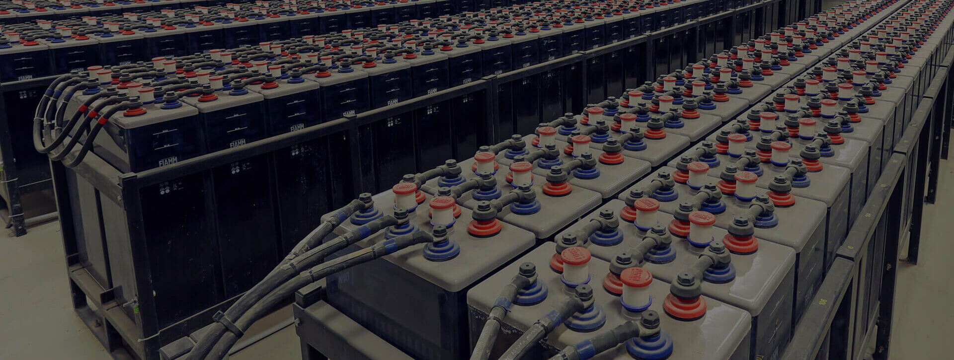 Rows of large industrial batteries, connected with black cables, fill the warehouse. The batteries have red and blue terminal caps and are arranged in multiple long lines.