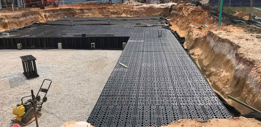 Construction site with a partially covered ground of black drainage cells laid over gravel. The area is surrounded by dirt and a deep excavation trench. Various construction equipment and materials are visible around the site.