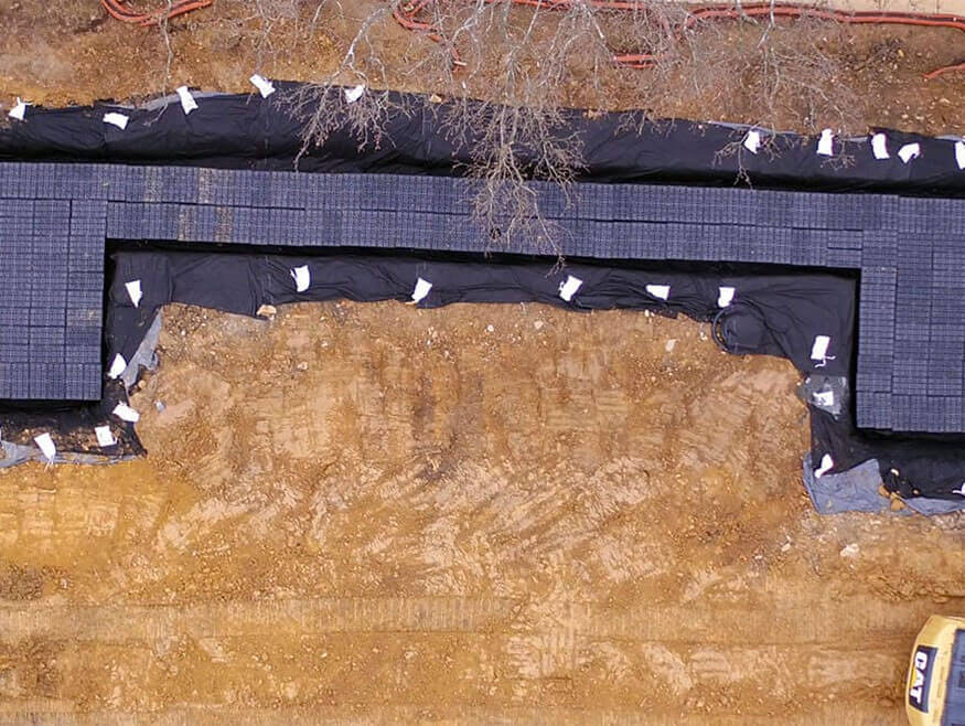 Vista aérea de una obra en construcción que presenta una zanja excavada revestida de tela negra y parcialmente rellena de cajas de plástico negro, diseñada para la gestión de las aguas pluviales.