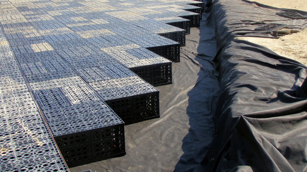 A construction site showing a partially completed installation of black plastic modular drainage cells arranged in a grid pattern over a black geotextile fabric. The grid extends diagonally, and the surrounding fabric is partially folded, highlighting the efficient stormwater management system being implemented.