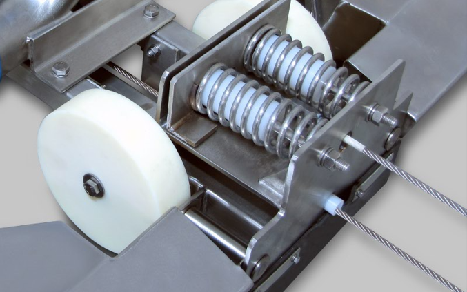 Close-up of a mechanical component featuring two large white gears, metal coils, and a tension system with a steel cable. The assembly is made of stainless steel and exhibits a precise industrial design.