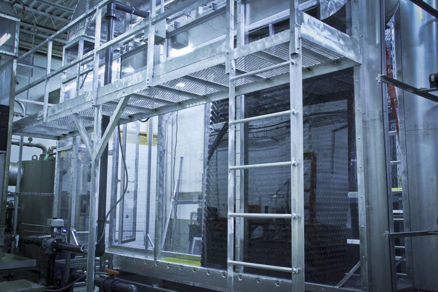 A complex industrial setup featuring metal frameworks and a ladder leading to a platform. The structure includes glass and metal components, with interconnected pipes and machinery visible in the background.