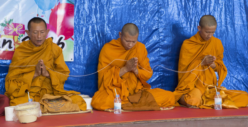 Tres monjes están sentados en fila sobre una estera, cada uno con su túnica. Sus manos están entrelazadas en un gesto de oración, unidas por un cordel blanco sostenido entre ellas.