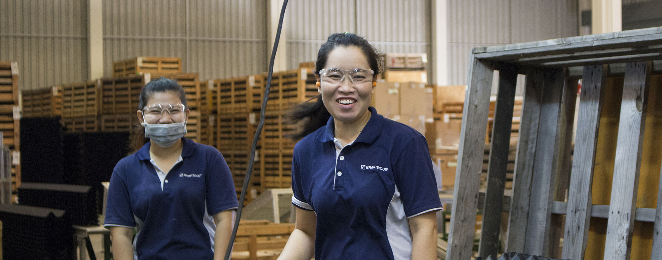 Two individuals in a warehouse setting are smiling at the camera. Both are wearing safety glasses and dark blue Brentwood shirts. One person has a face mask pulled down, and there are stacks of wooden pallets in the background.