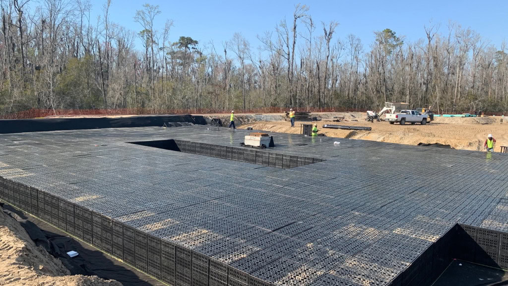 A construction site with a large metal grid structure covering the ground, surrounded by trees. Several workers in safety gear are present, and construction equipment is visible in the background.