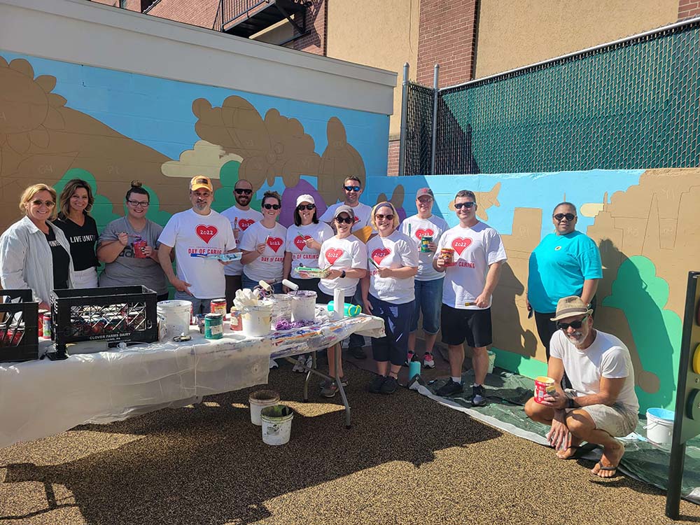 A group of people, some wearing t-shirts with hearts and text, pose in front of a colorful mural in progress. Paint cans and brushes are on a table in front of them. The setting appears to be an outdoor space on a sunny day, showcasing the vibrant culture of the community