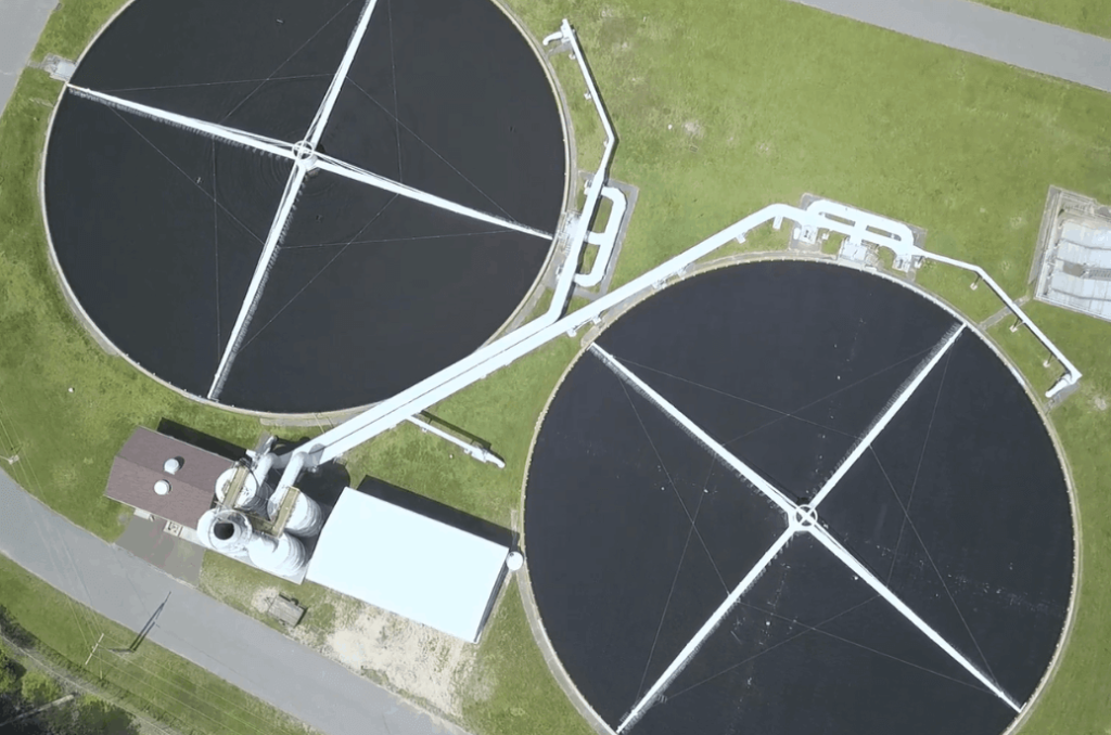 Aerial view of two large circular water treatment tanks with several pipes connected to them, situated on a grassy area. Small buildings are adjacent to the tanks.