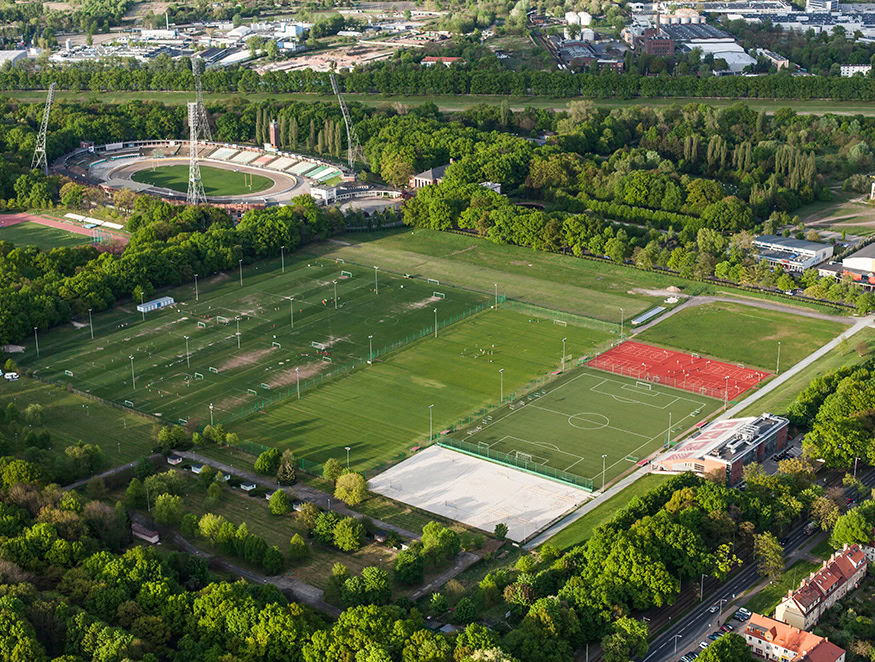 Aerial view of a sports complex featuring multiple sports fields, including green soccer pitches, a red athletics track, tennis courts, and an adjacent stadium. The complex incorporates stormwater management systems.