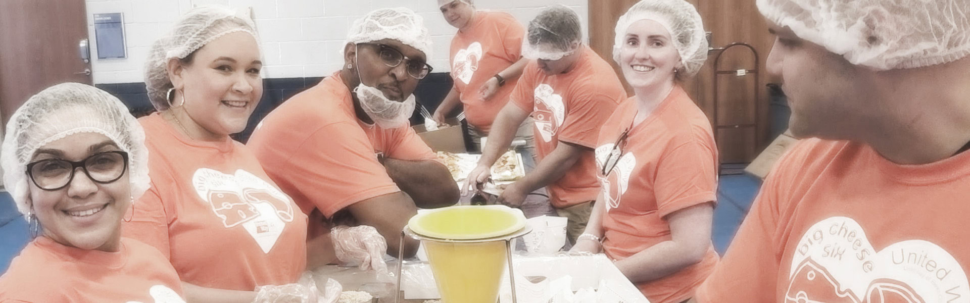 A group of people wearing hair nets and matching orange t-shirts sit and stand around a table, engaged in meal preparation. The room appears to be a community center or kitchen. They appear cheerful and collaborative.