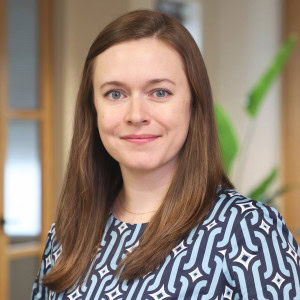 A woman with long brown hair is smiling at the camera in an indoor setting. Caitlin Banta is wearing a blue and white patterned top, and there is a blurred background with a wooden door and some green plants.