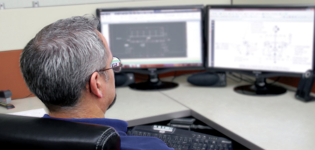 Una persona con pelo corto y canoso y gafas está sentada en un escritorio trabajando en dos monitores de ordenador. Las pantallas muestran dibujos técnicos y diagramas relacionados con servicios de ingeniería.