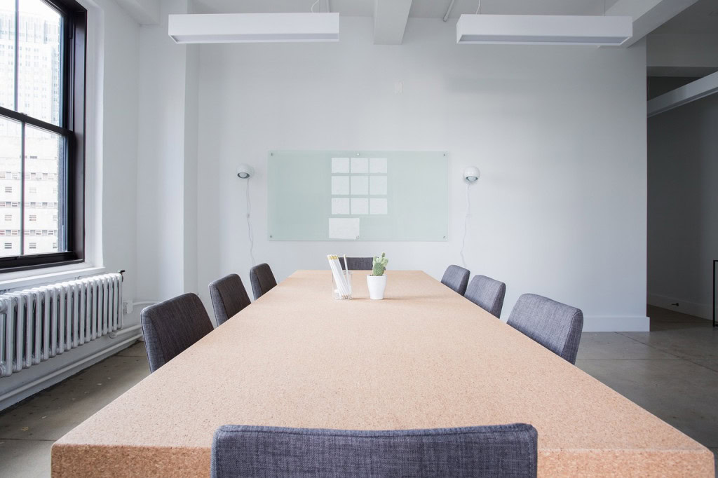 A modern conference room with a long wooden table surrounded by eight gray chairs. The table has a small desk organizer with pens and a potted cactus.