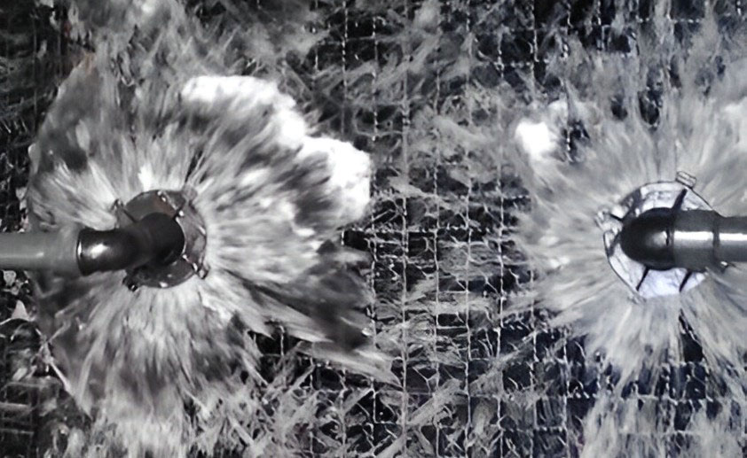 Two circular water jets spraying water outward in a pattern against a dark, grated surface. The nozzles create symmetrical, splashing water bursts.