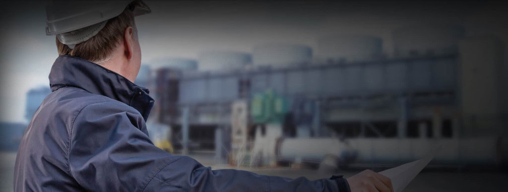 A worker wearing a helmet and a blue jacket faces away, looking at industrial cooling towers in the distance. He holds a sheet of paper, possibly plans or blueprints, in his left hand.