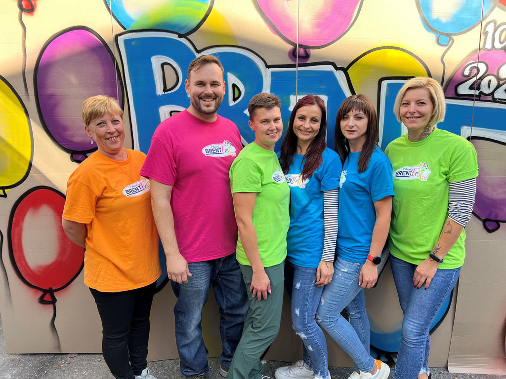 A group of six smiling people stand together in front of a colorful balloon-themed backdrop. They wear bright t-shirts in various colors, each with a "BrentFest" logo.