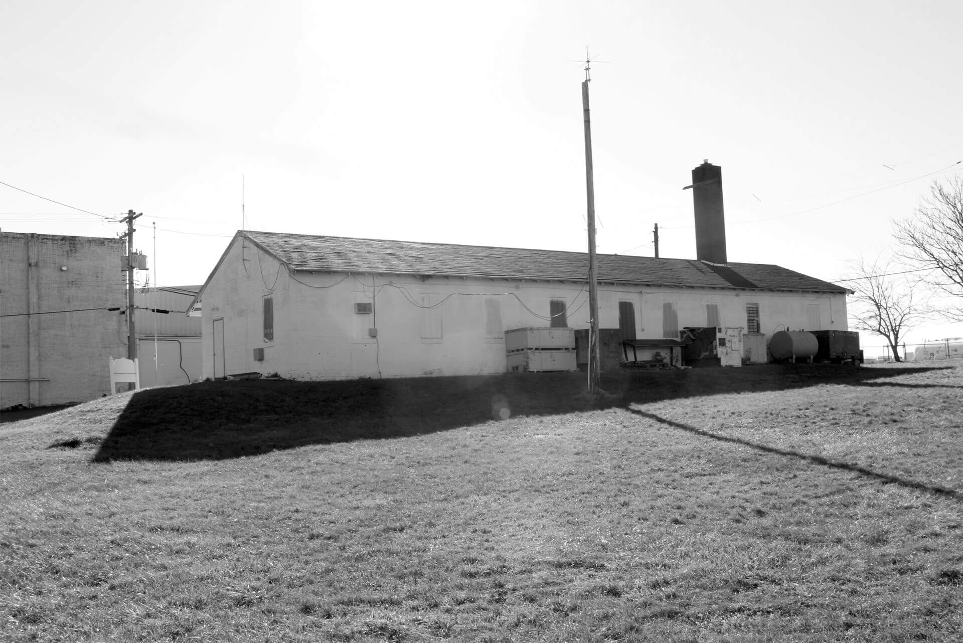 A black-and-white photo of a long, rectangular, single-story building with a slanted roof. Utility poles, wires, and equipment surround the structure.