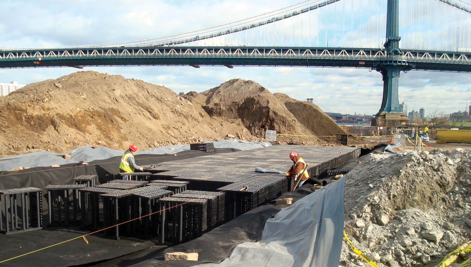 Construction workers in safety gear are working on a site with large piles of dirt and construction materials. There is a bridge in the background spanning across the image.