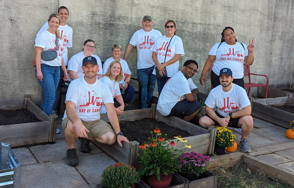 Un grupo de once personas con camisetas blancas del "Día de la solidaridad" posan juntas en un jardín con parterres en Reading, Pensilvania. Algunos están en cuclillas o sentados en los parterres del jardín, mientras que otros permanecen de pie detrás de ellos. Hay coloridas flores en macetas y calabazas alrededor de la zona ajardinada.