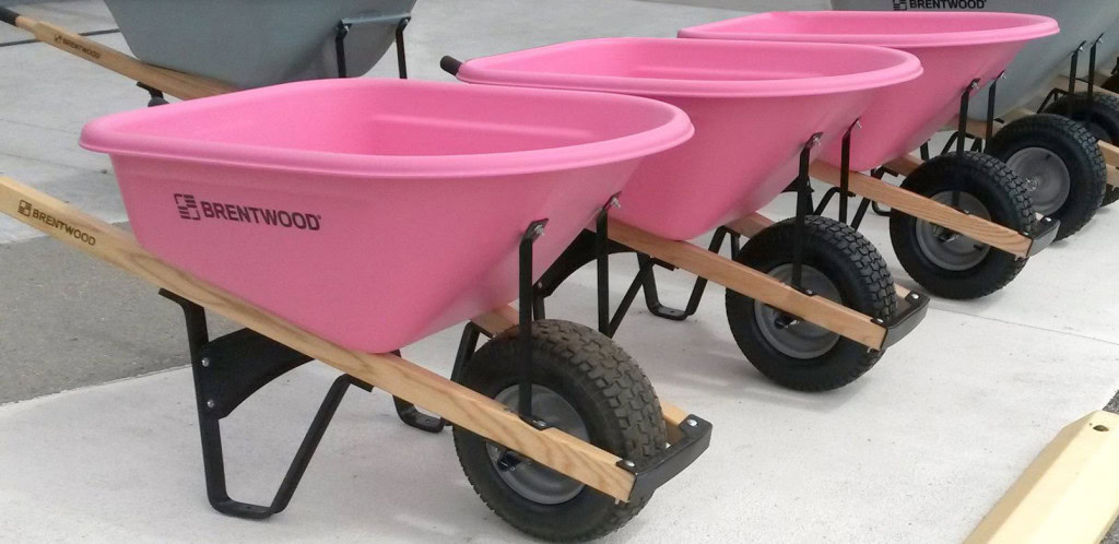 A row of light pink wheelbarrows with black frames and wooden handles, each featuring the brand name "Brentwood," are lined up on a concrete surface.