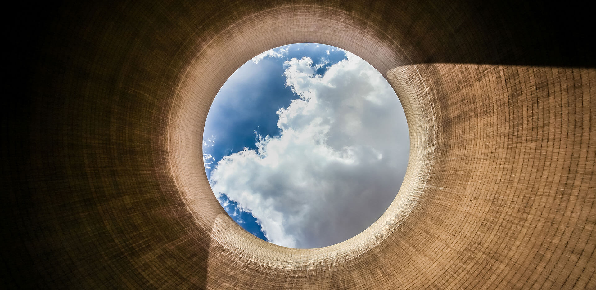 A view from the bottom of an industrial cooling tower looking up at a circular opening showing a portion of a blue sky with white clouds.