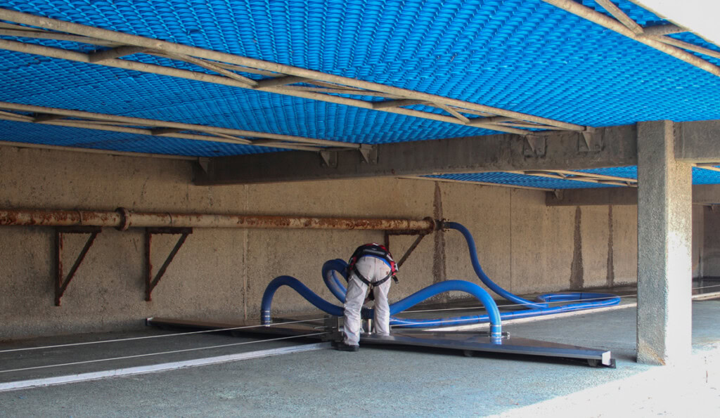 Una persona trabaja en un gran espacio industrial, manipulando mangueras azules conectadas a un sistema SedVac™. El techo está forrado de malla azul y unas robustas vigas sostienen la estructura.