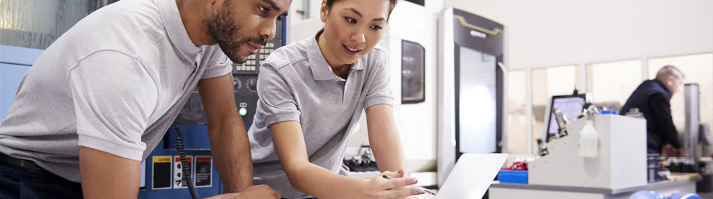 Two workers, a man and a woman, are looking at a tablet while standing near industrial machinery. They appear to be discussing something on the screen. Both are wearing gray shirts.