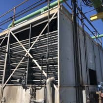 An industrial cooling tower with visible metal framework and large horizontal slats is equipped with several pipes, valves, and fills