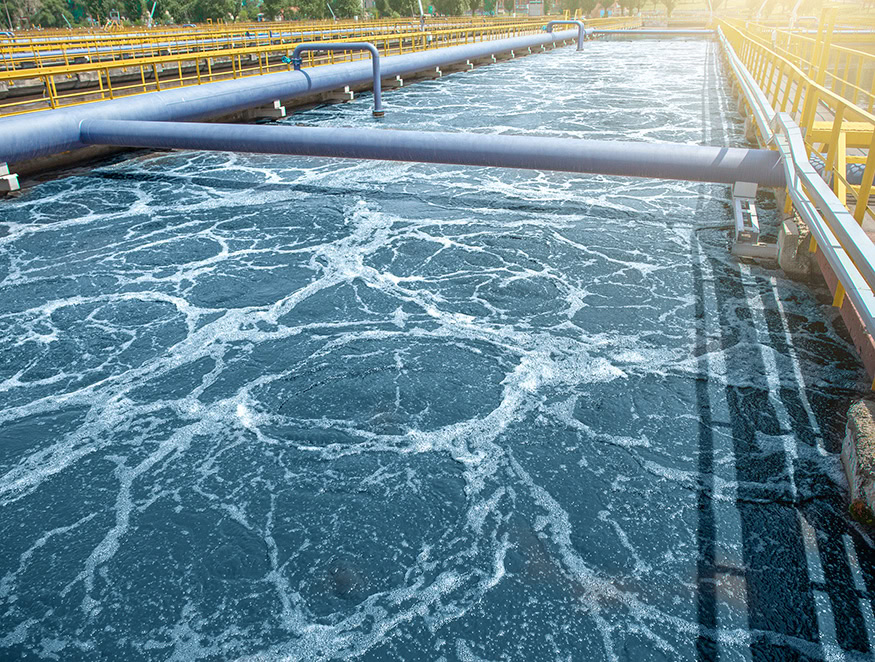 A large wastewater treatment facility with multiple pools of agitated, foamy water. Blue pipes run across the pools and sunlight can be seen reflecting on the water and pipes.