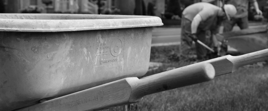 Black and white image showing a close-up of a wheelbarrow with two wooden handles in focus. In the background, blurred construction workers are seen digging with shovels