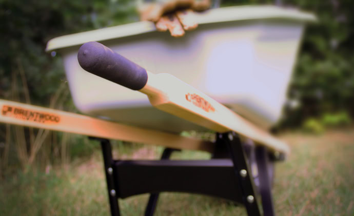 Close-up image of a Brentwood wheelbarrow handle with a blurred background. The handle is wooden with a black grip, and the wheelbarrow is light-colored. The background features greenery.