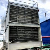 A large industrial cooling tower fills the view against a partly cloudy sky. The rectangular structure features metal grates on the front and a set of stairs or ladders on the side.