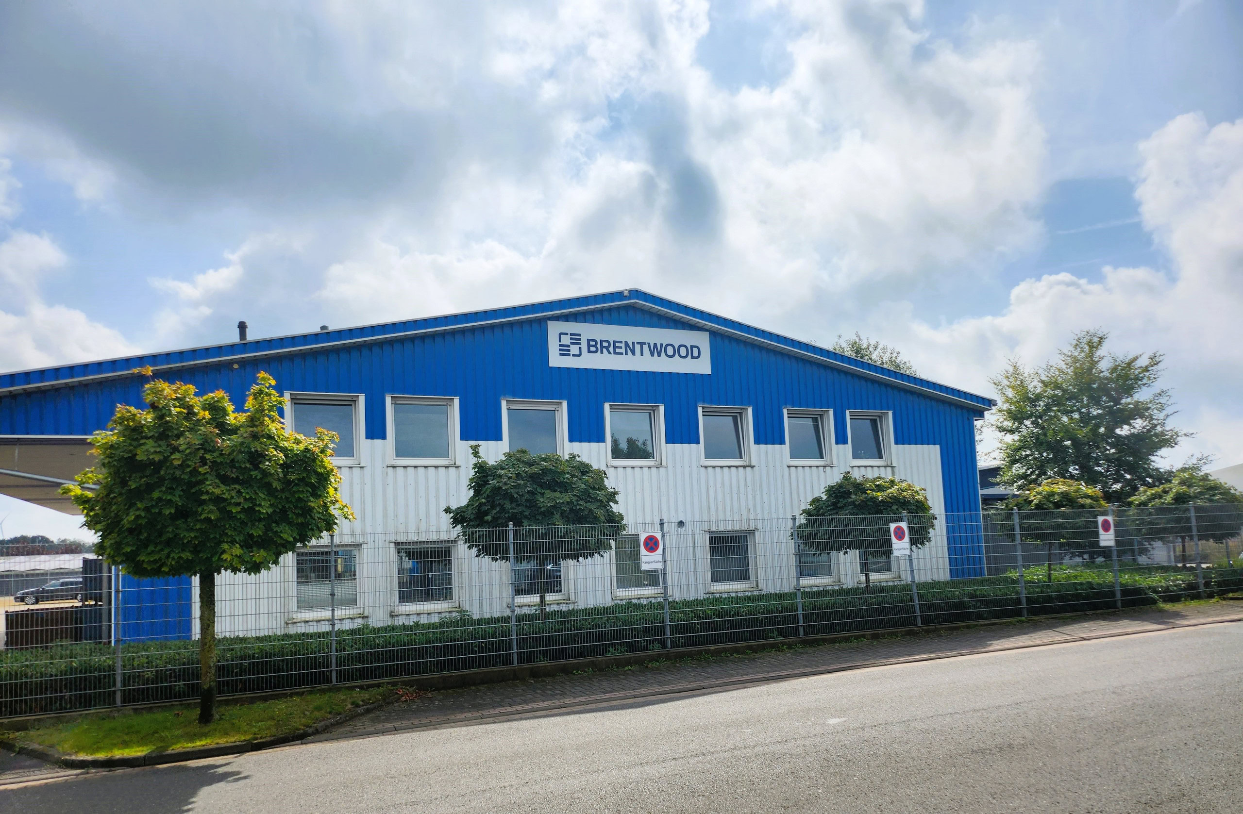 A blue and white industrial building with the name "Brentwood" on the facade, behind a fenced area. The building has several windows and is surrounded by trimmed greenery.