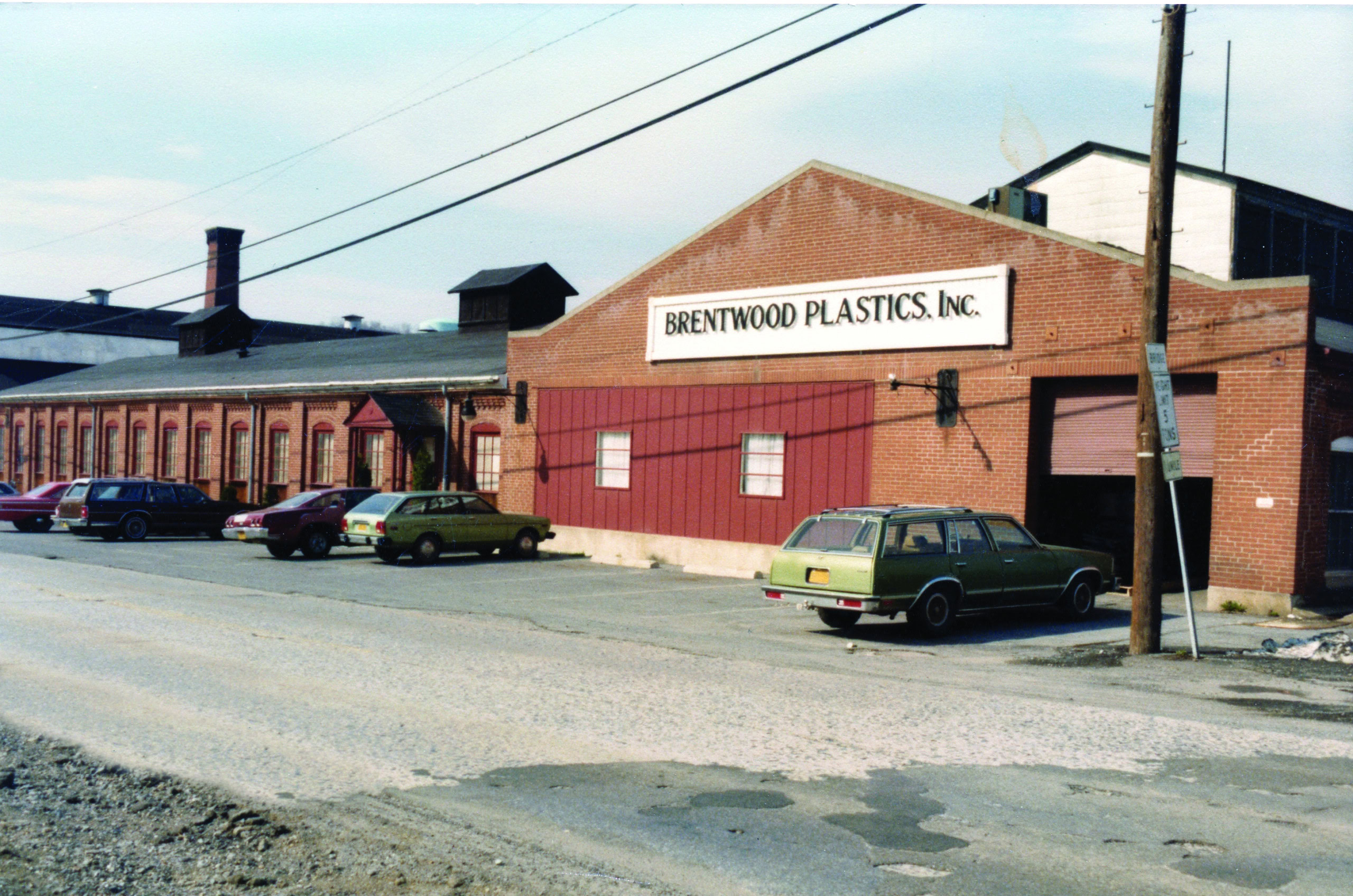 Un almacén de ladrillo de una planta con un letrero blanco rectangular en la parte superior en el que se lee "Brentwood Plastics, Inc.". Hay varios coches aparcados a lo largo de la fachada y una puerta de garaje abierta en el lado derecho.