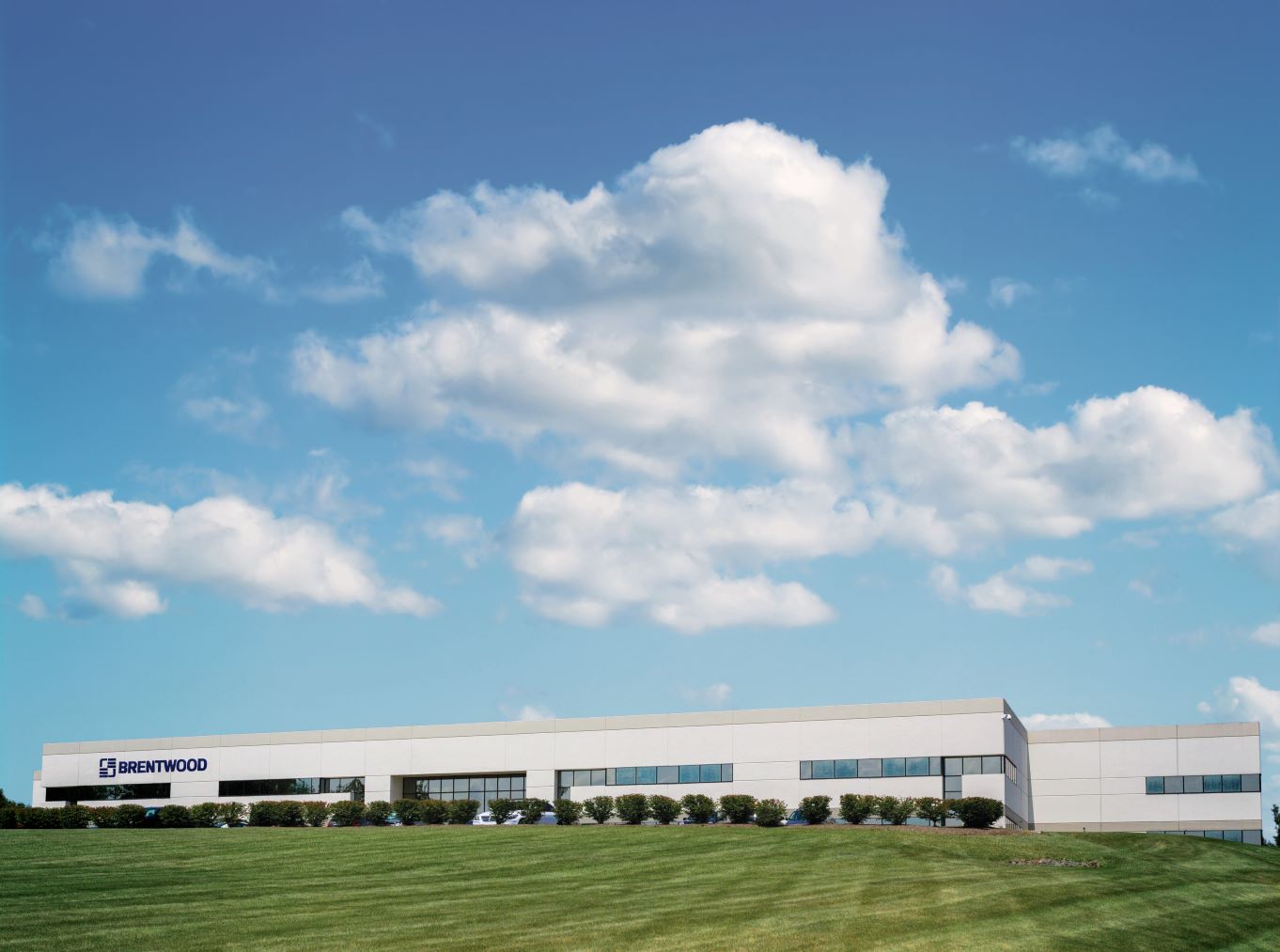 A modern, single-story office building with a white exterior sits on a well-manicured, grassy hill against a backdrop of a blue sky with scattered, fluffy white clouds.