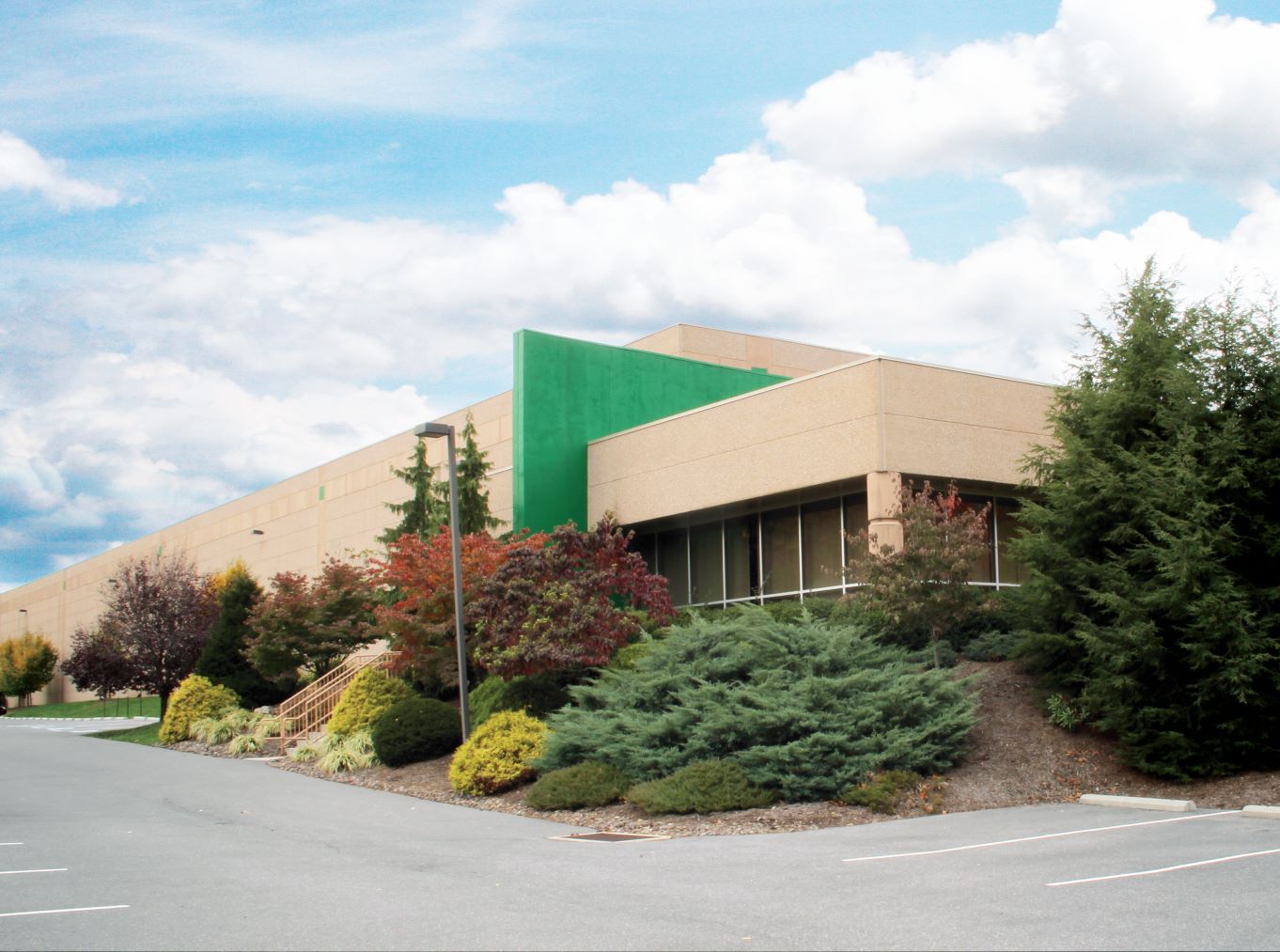 A large beige industrial building with a green accent wall is surrounded by a variety of trees and shrubs. The front of the building features large windows, and the sky above is blue with scattered clouds.