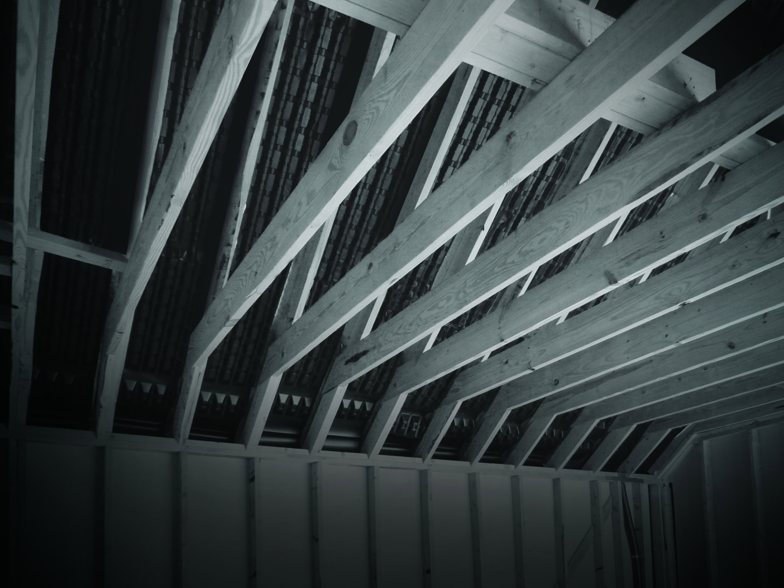 A dimly lit image of a partially constructed roof in a building. The wooden beams supporting the roof are visible, with an exposed structure showcasing the framework and rafters.