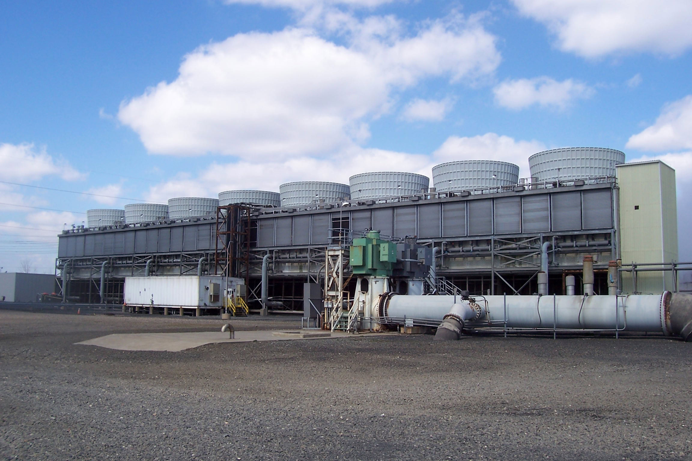 Una gran instalación industrial de torres de refrigeración bajo un cielo azul con nubes dispersas. La estructura presenta múltiples unidades de refrigeración cilíndricas sobre un edificio rectangular, con tuberías.