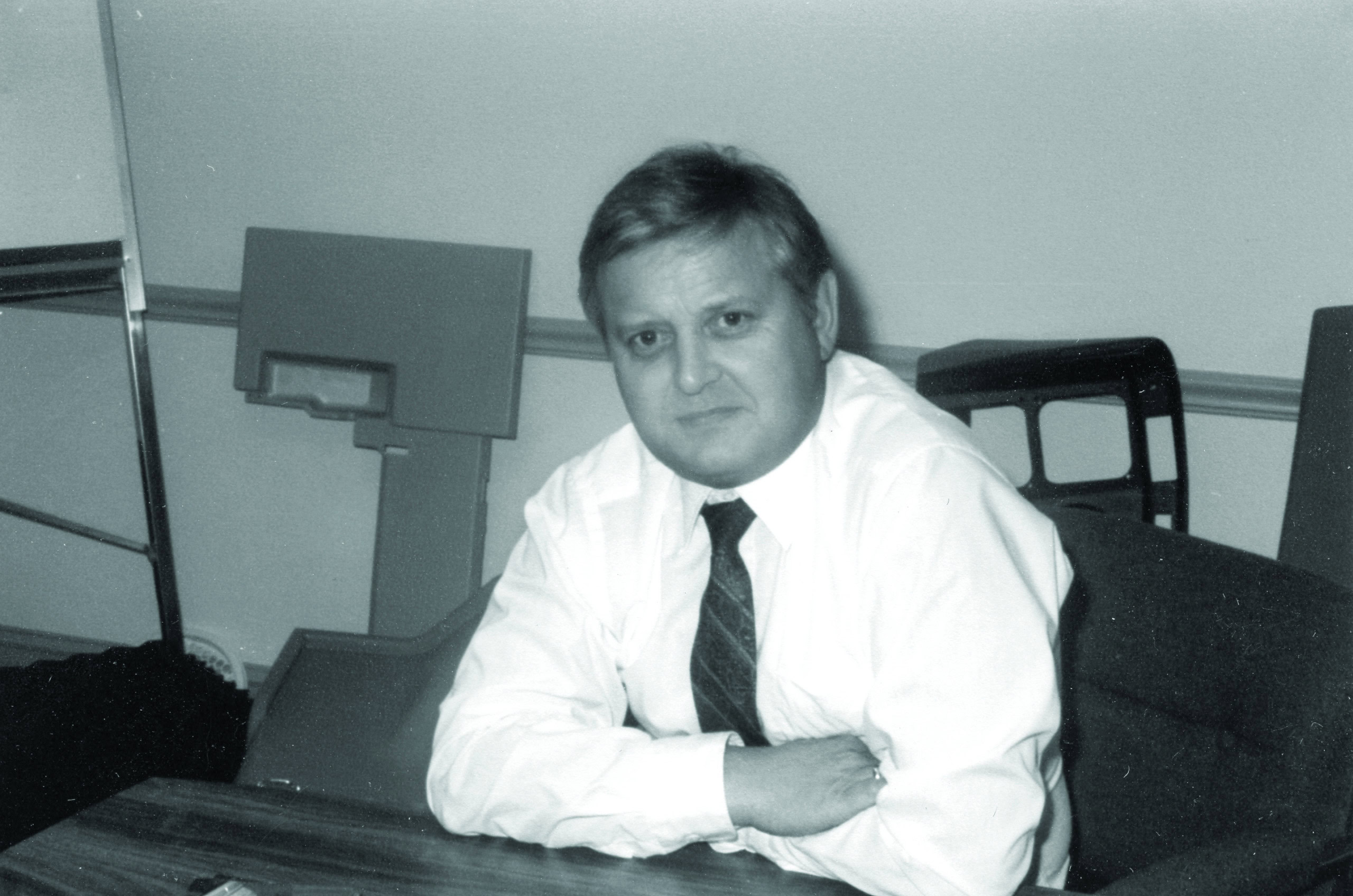 A man with short hair and a tie sits at a wooden table, leaning slightly forward as he looks at the camera with a neutral expression. The office setting in the background features chairs and partitions, evoking an aura of timeless history. The image is in black and white.