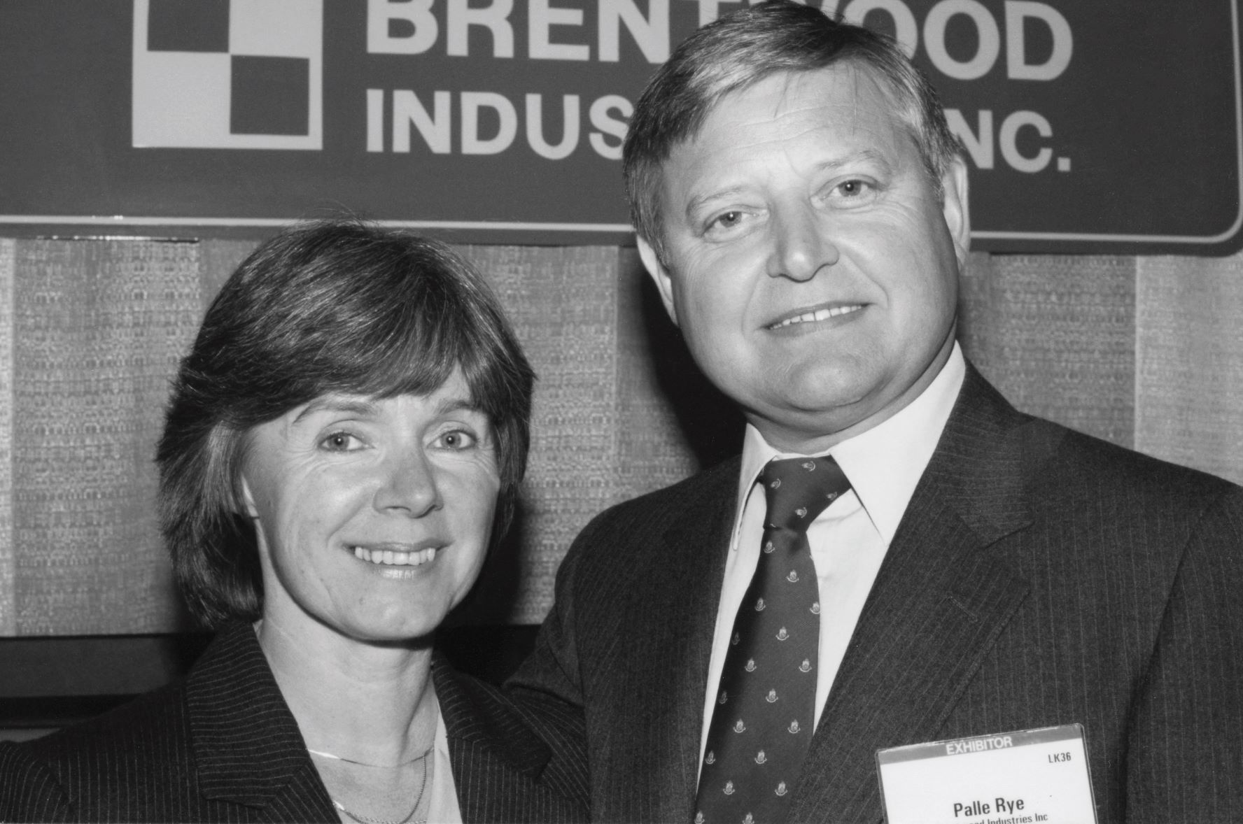 A black and white photo shows a man and a woman smiling at the camera, dressed in formal business attire. Behind them is a sign that reads "Brentwood Industries, Inc.," The man has a name tag that says "Palle Rye.