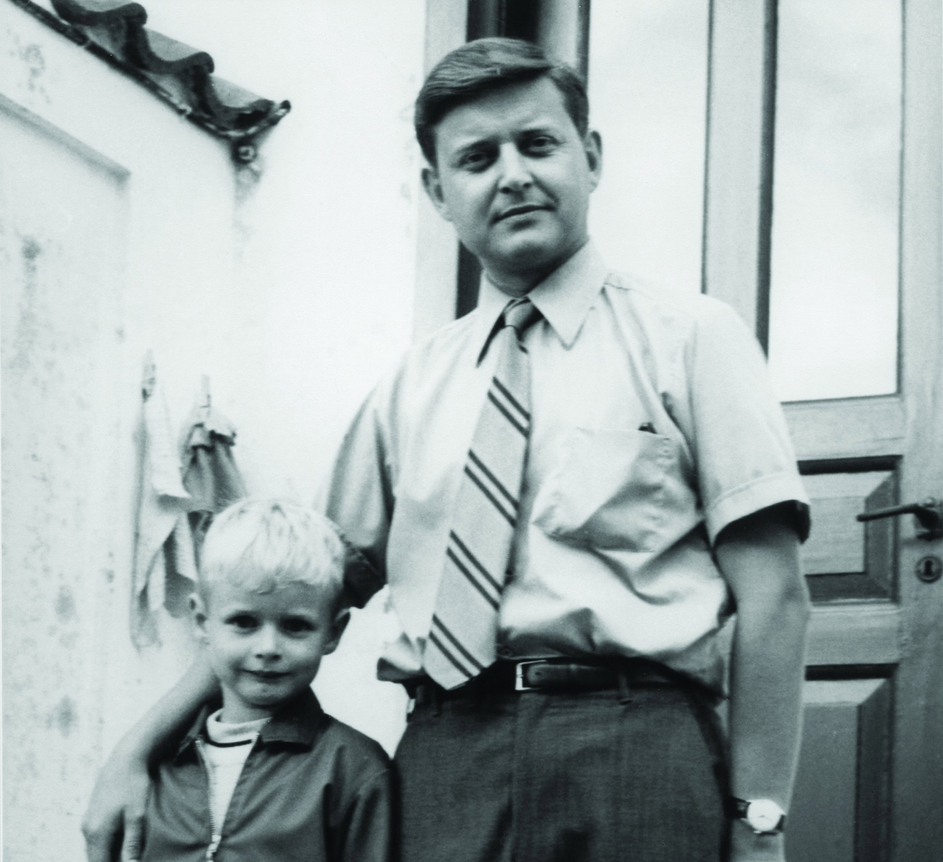 Fotografía en blanco y negro de un hombre y un niño pequeño. El hombre, vestido con camisa de manga corta, corbata y pantalón, rodea con el brazo al niño, que lleva una chaqueta con cremallera. Ambos están de pie delante de una puerta.