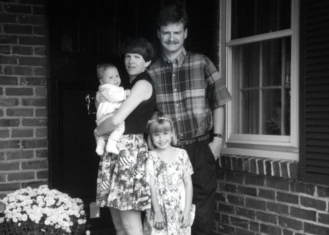 A black-and-white photo captures a moment in history: a family of four standing in front of a brick house. The mother cradles a baby, while the father stands beside them, hand in pocket. A young girl grins next to them.