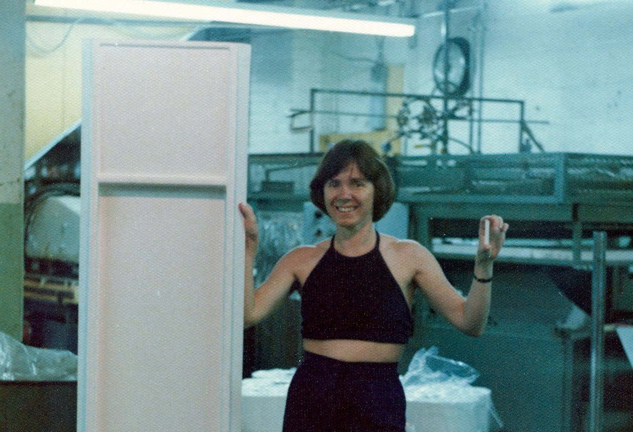 A woman with short brown hair, wearing a dark top and pants, smiles and gestures with both hands in an industrial setting. She stands beside a tall, narrow, rectangular object.