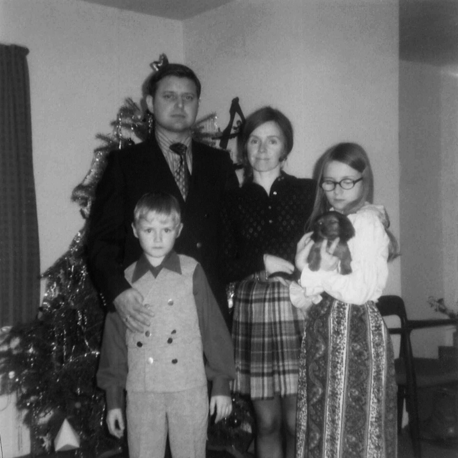 Fotografía en blanco y negro de una familia de pie delante de un árbol de Navidad decorado. La familia está formada por un hombre, una mujer, un niño pequeño y una niña que sostiene un perro pequeño. Todos están vestidos con atuendos formales apropiados para las fiestas.