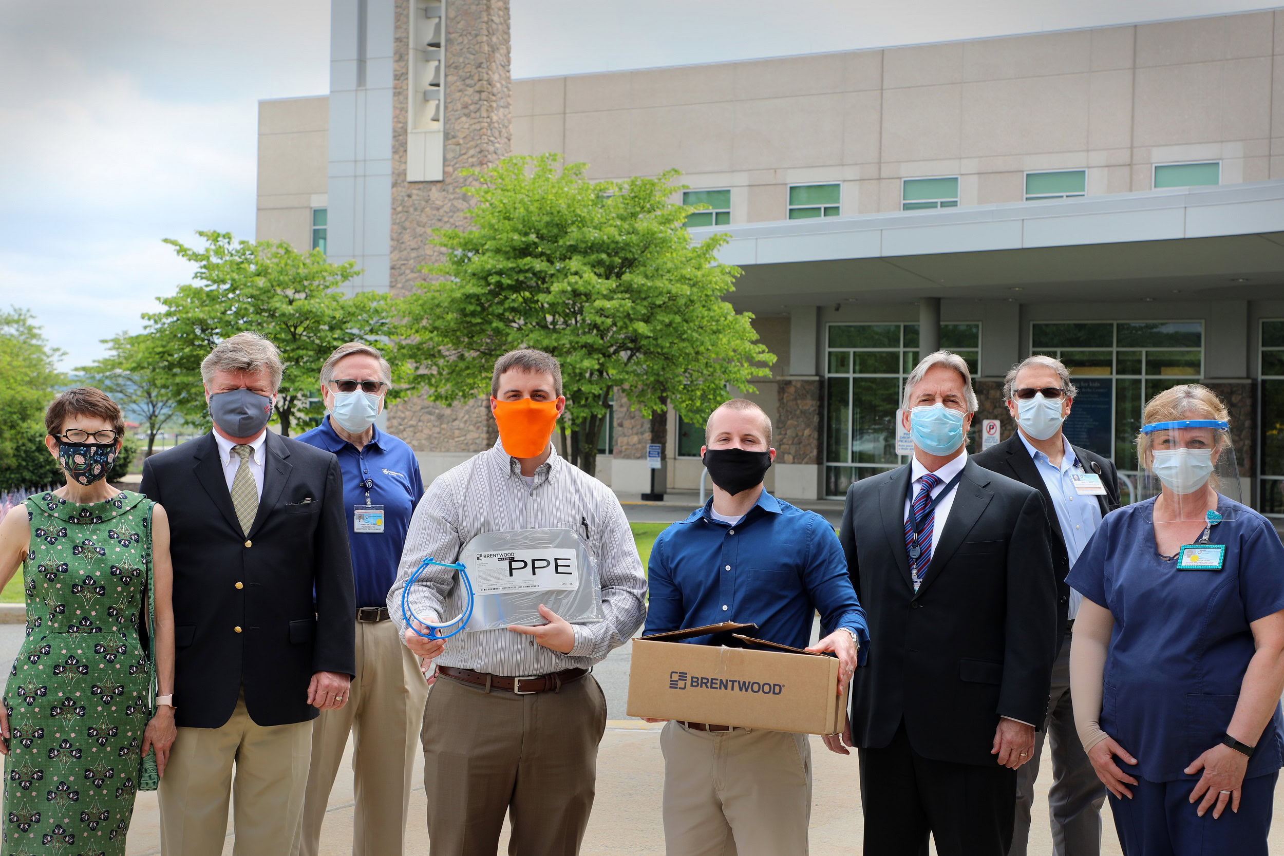 A group of eight individuals wearing masks stands outside a building with large windows. Two people in the center hold boxes labeled "PPE" and "Brentwood."