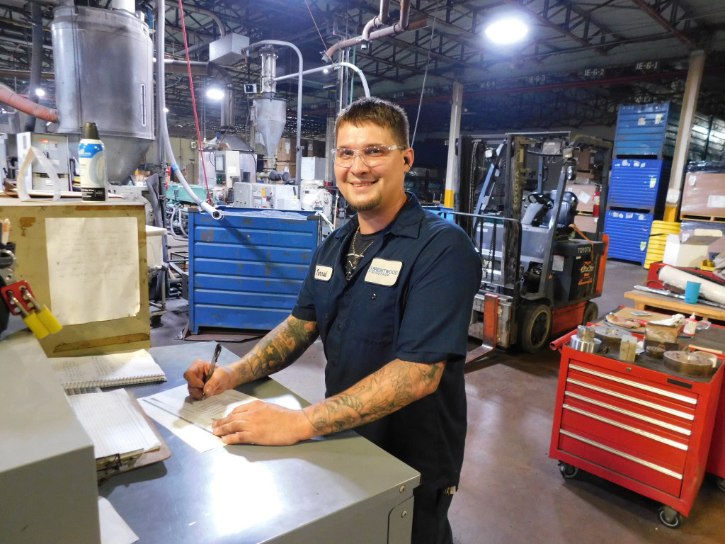 A man in a blue Brentwood uniform and safety glasses is smiling and writing on a clipboard in an industrial setting. Behind him are various machines, cabinets, and a forklift in a warehouse.