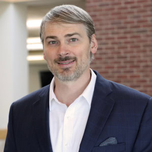 Clinton McCorkle, a man with light hair and a beard, is smiling at the camera. He is wearing a dark blue suit jacket, a white shirt, and a pocket square.