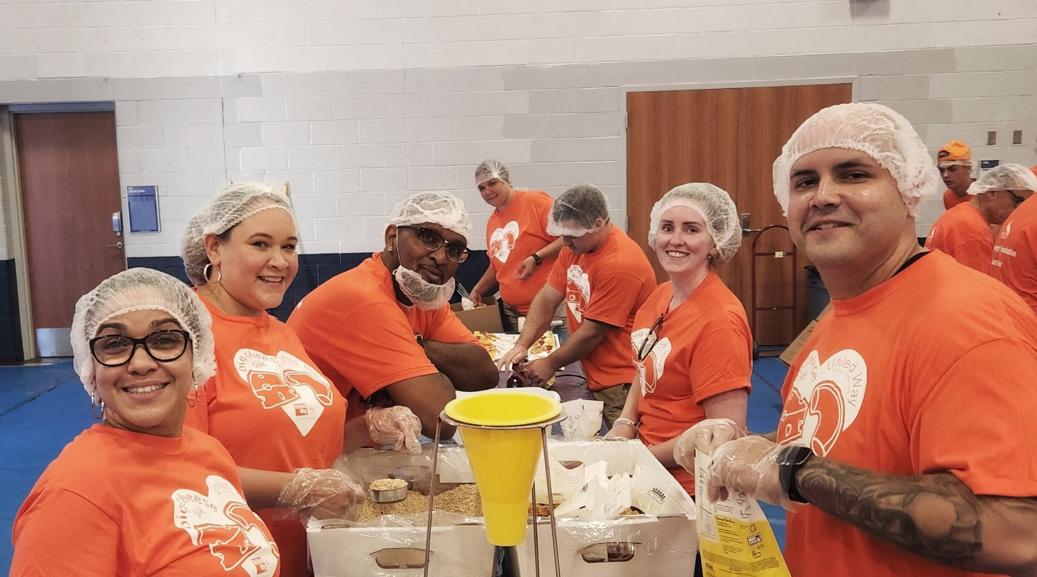 A group of volunteers wearing orange T-shirts and hairnets stand around tables in a gymnasium, smiling and preparing food packages. They are engaged in a community service event.