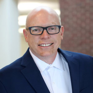 Dennis Reed, a bald man with glasses, smiles warmly at the camera. He is wearing a navy blue blazer over a white shirt. The background is softly blurred.