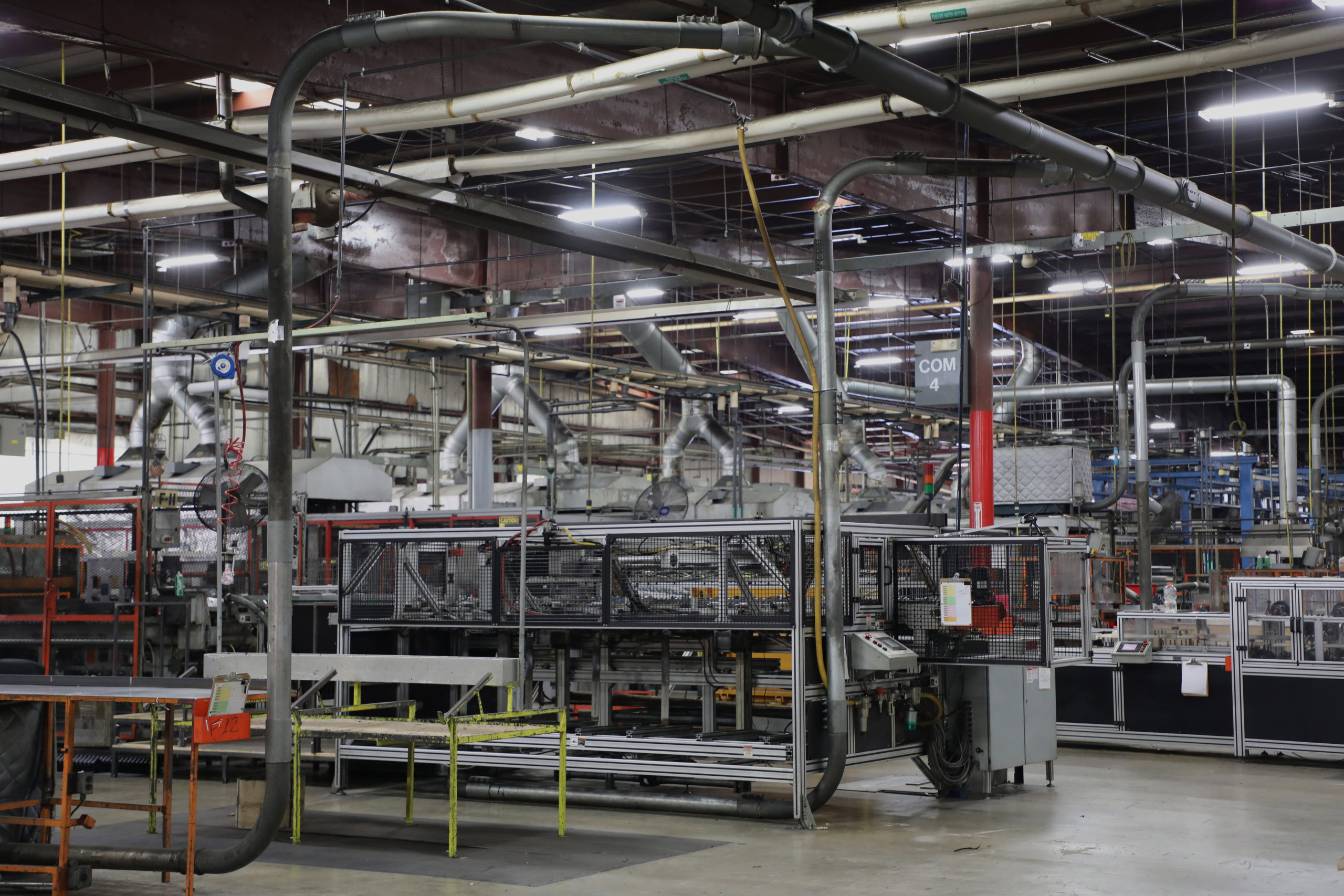 A wide shot of a large industrial manufacturing facility with various machinery, pipes, and equipment. Conveyor belts and workstations are visible under bright overhead lighting.