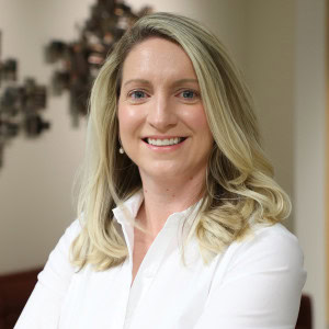 Kristin Kohler, a woman with long blonde hair wearing a white blouse, stands with her arms crossed.